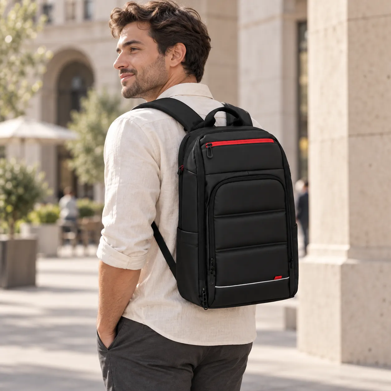 Entspannter Mann in hellem Leinenhemd trägt den schwarzen Laptop Rucksack mit rotem Reissverschluss auf dem Rücken in einer sonnigen Altstadt